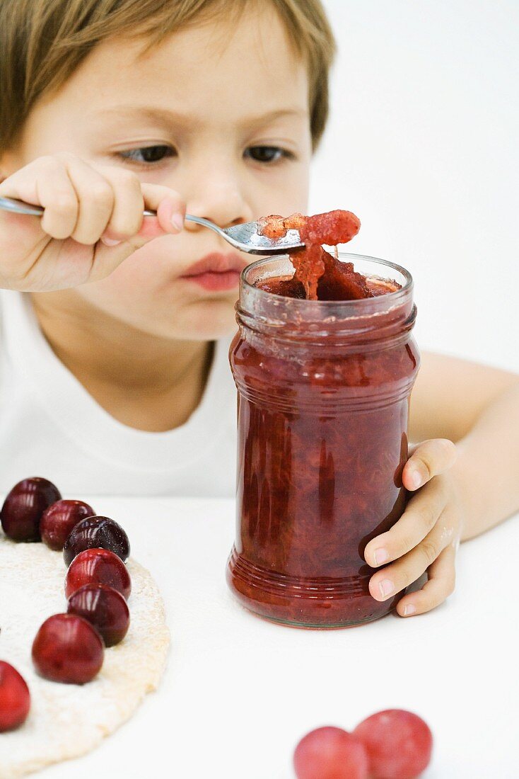 Little boy scooping jam out of jar with spoon, tart and cherries on table in front of him