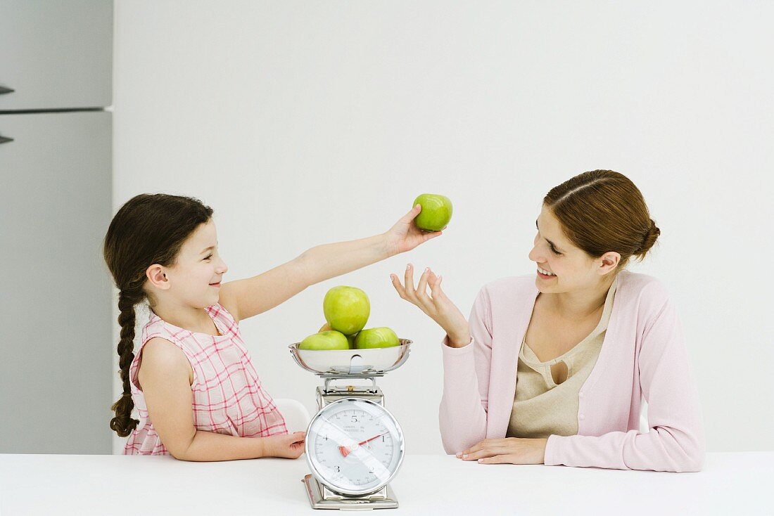 Mother and daughter weighing apples on scale, smiling at each other, girl holding up one apple