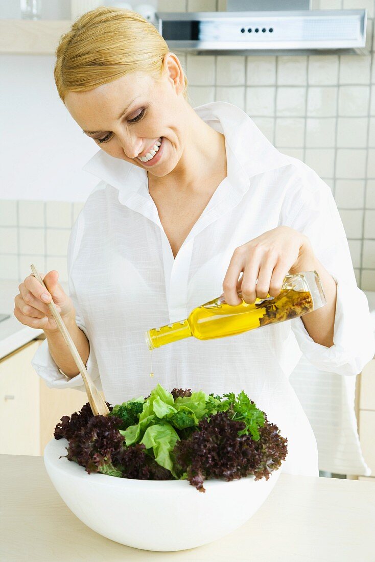 Woman in kitchen, pouring olive oil on large salad