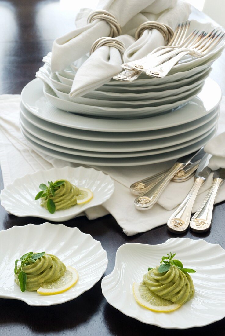 Muschelförmige Schalen mit Avocadocreme vor Tellerstapel und Stoffservietten mit Silberring