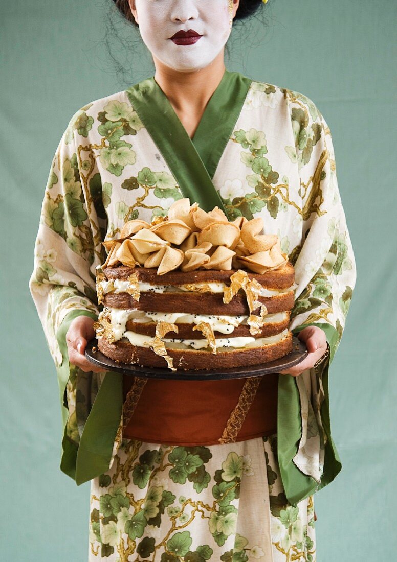 An Asian woman holding a celebration cake decorated with gold leaf and fortune cookies