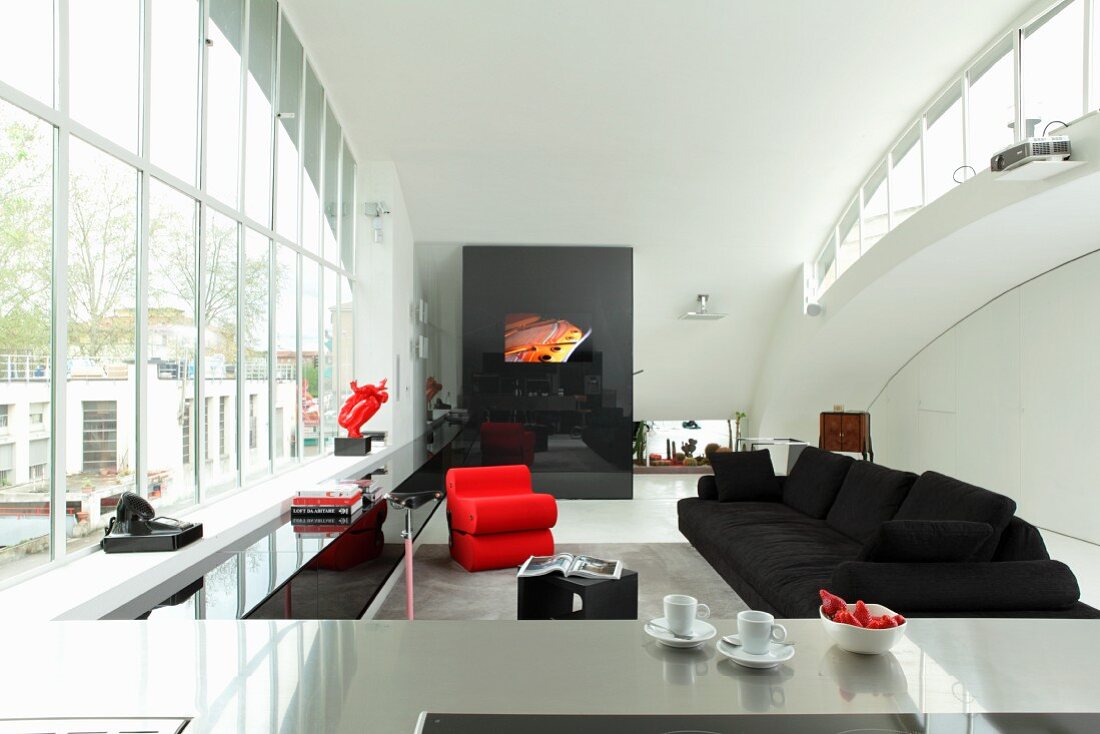 View across stainless steel kitchen counter to open-plan interior with black sofa and red designer armchair next to huge window