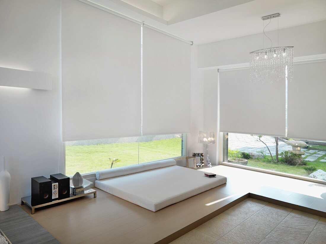 Minimalistisches Schlafzimmer mit podestartigem Holzeinbau und Liegepolster vor Fenster mit geschlossenem Rollo