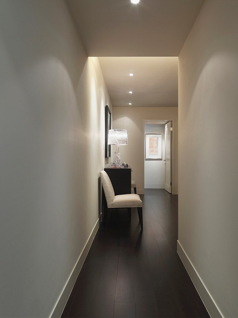 Upholstered chair next to chest of drawers in narrow corridor with recessed spotlights