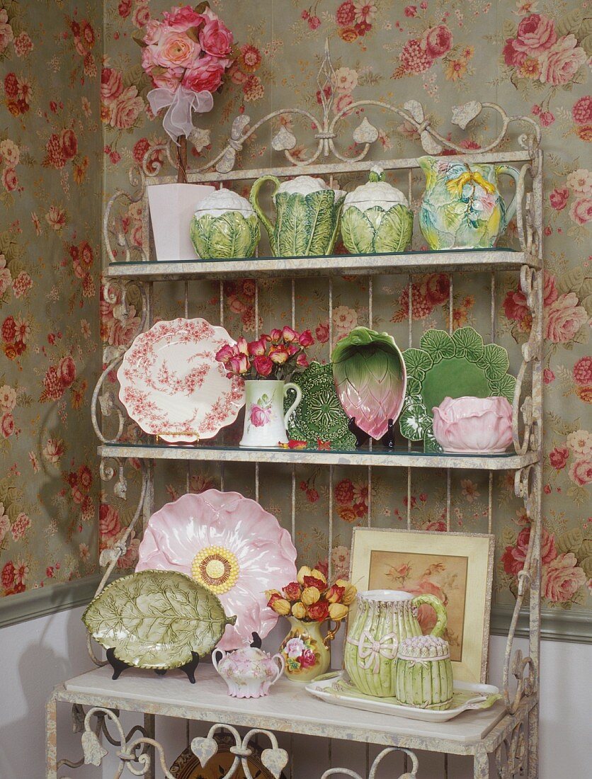 Collection of unusual crockery on artistic shabby chic metal shelving against rose-patterned wallpaper