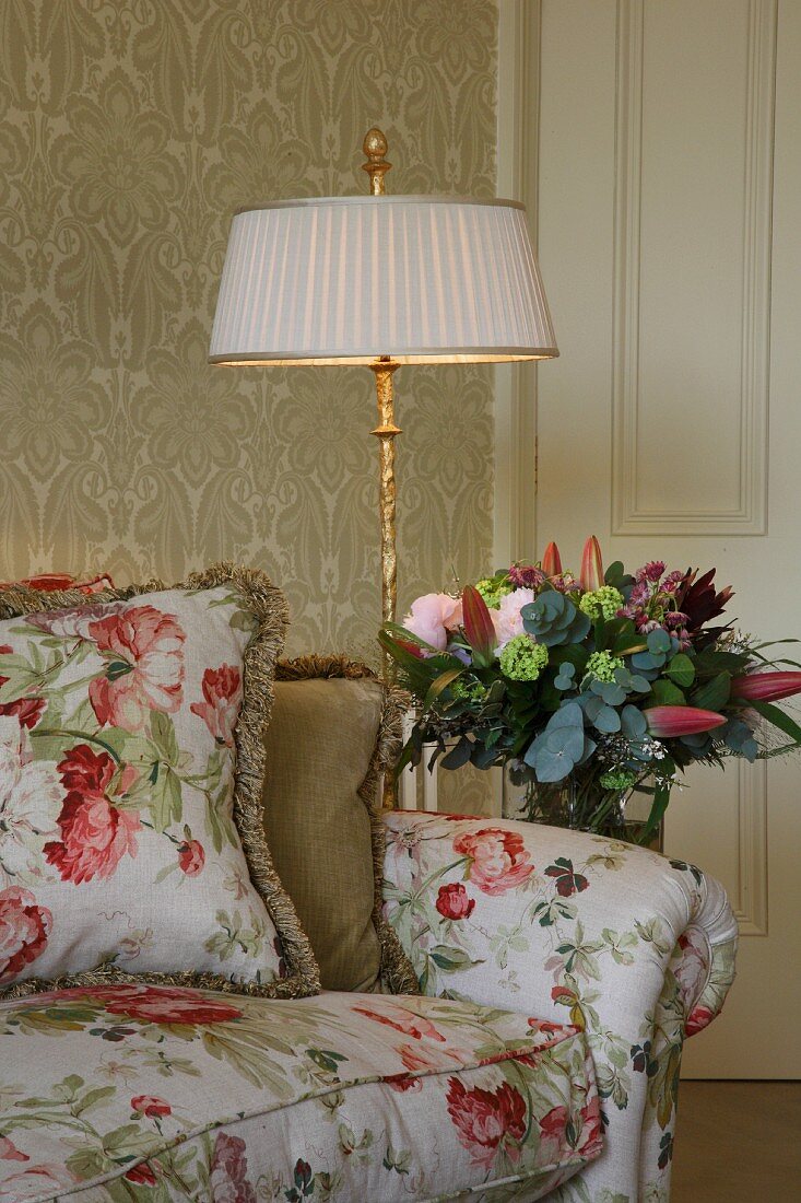 Floral sofa with cushions in same fabric next to standard lamp and bouquet in semi-darkness
