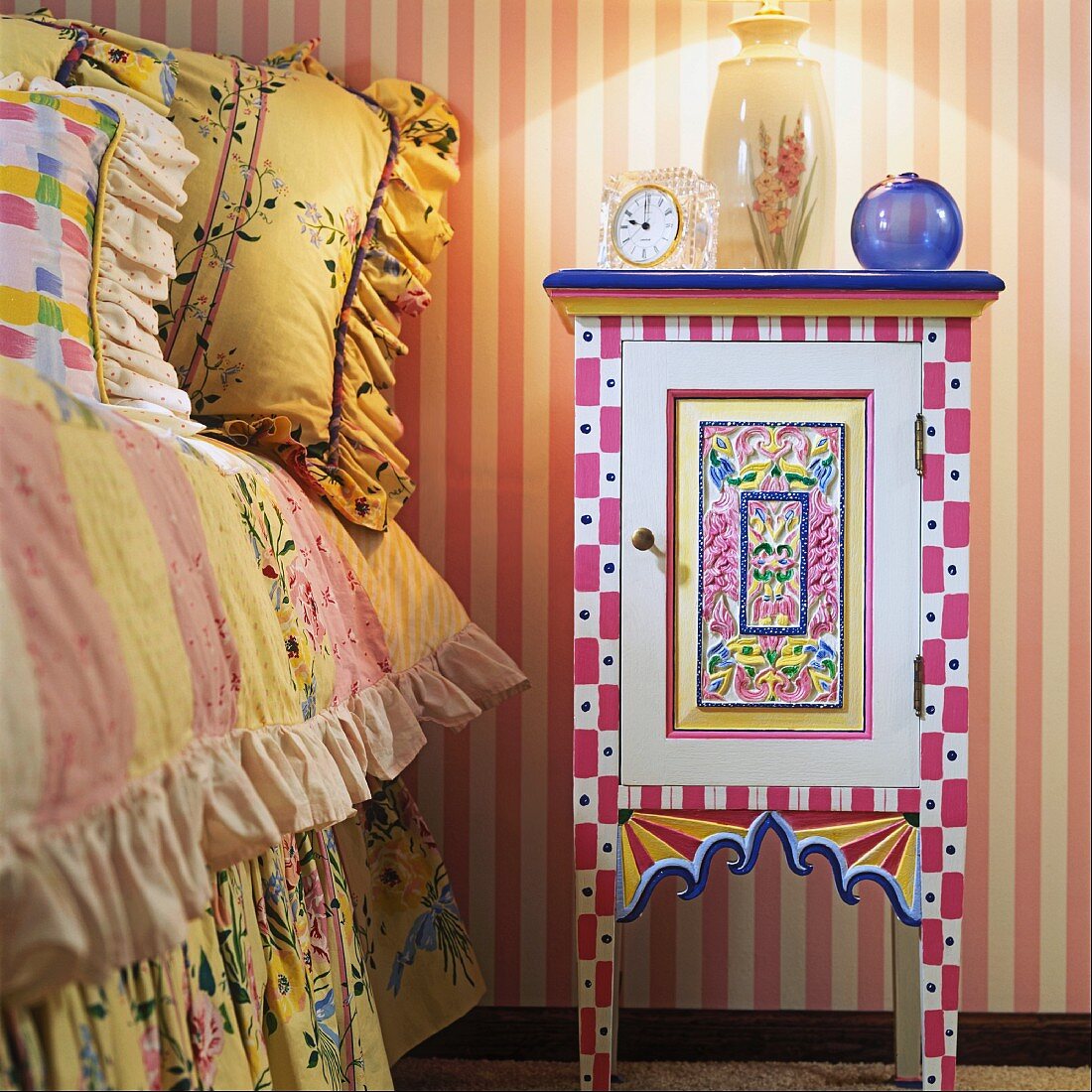 Hand-painted, colourful bedside cabinet next to child's bed with pretty cushions and ruffled bedspread; pink and white striped wallpaper