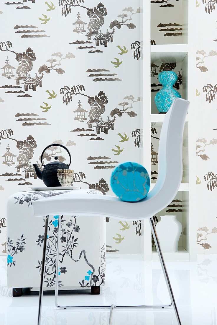 Light blue bolster on white modern chair in front of tea service, patterned wallpaper and vases on shelves