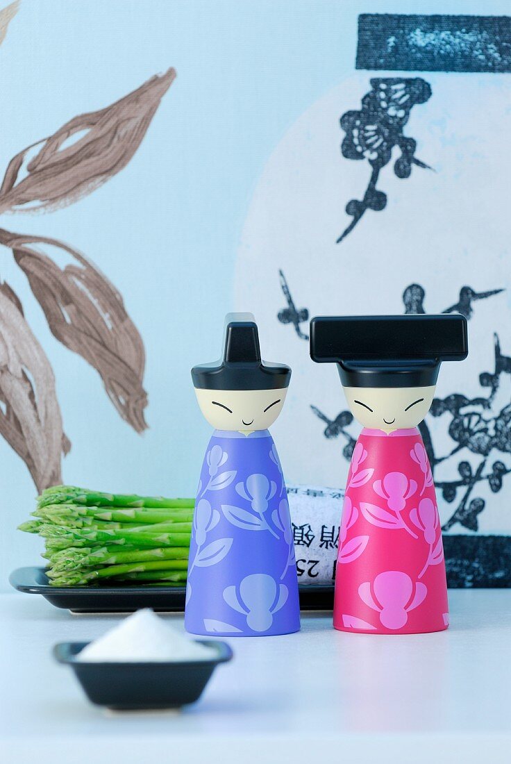 Japanese-style, painted wooden figurines on table with salt cellar and plate of asparagus against patterned wallpaper