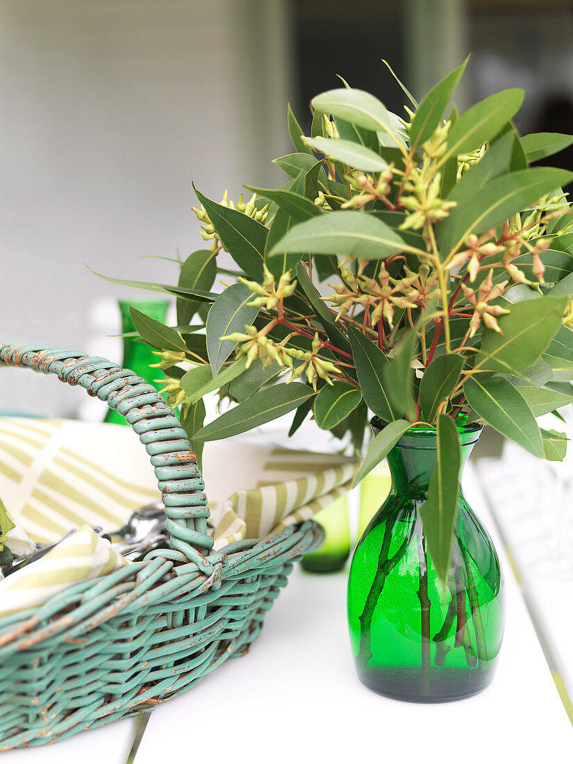 Vase of eucalyptus buds on white table next to basket of linen napkins