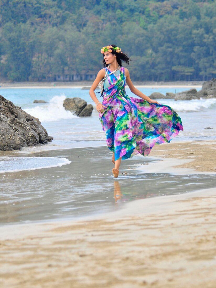 A woman wearing brightly patterned dress and wreath of flowers on her head running along a beach