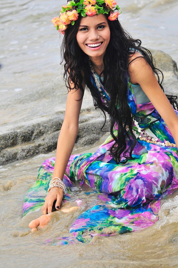 A woman sitting on a beach wearing brightly patterned stress and a wreath of flowers on her head