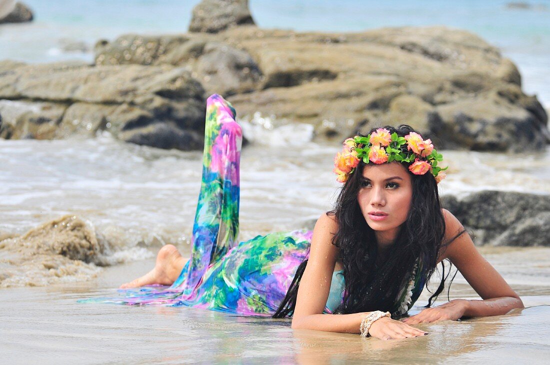 A woman lying on a beach wearing brightly patterned dress and wreath of flowers on her head