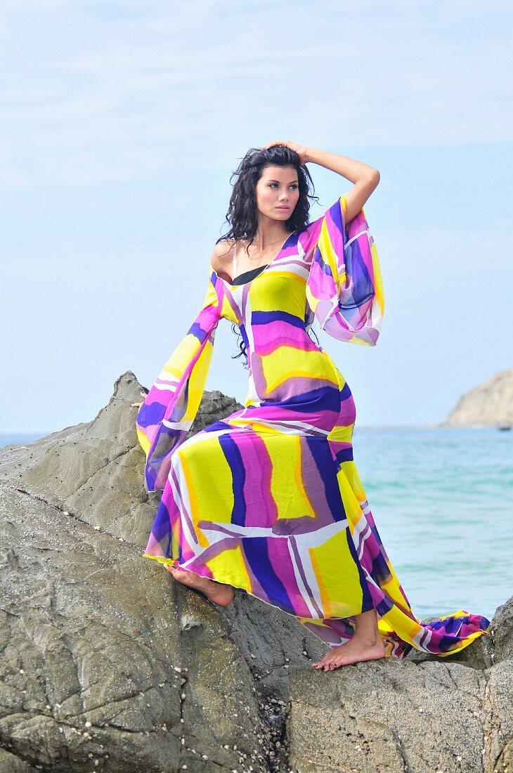 A woman on rocks on a beach wearing brightly patterned dress