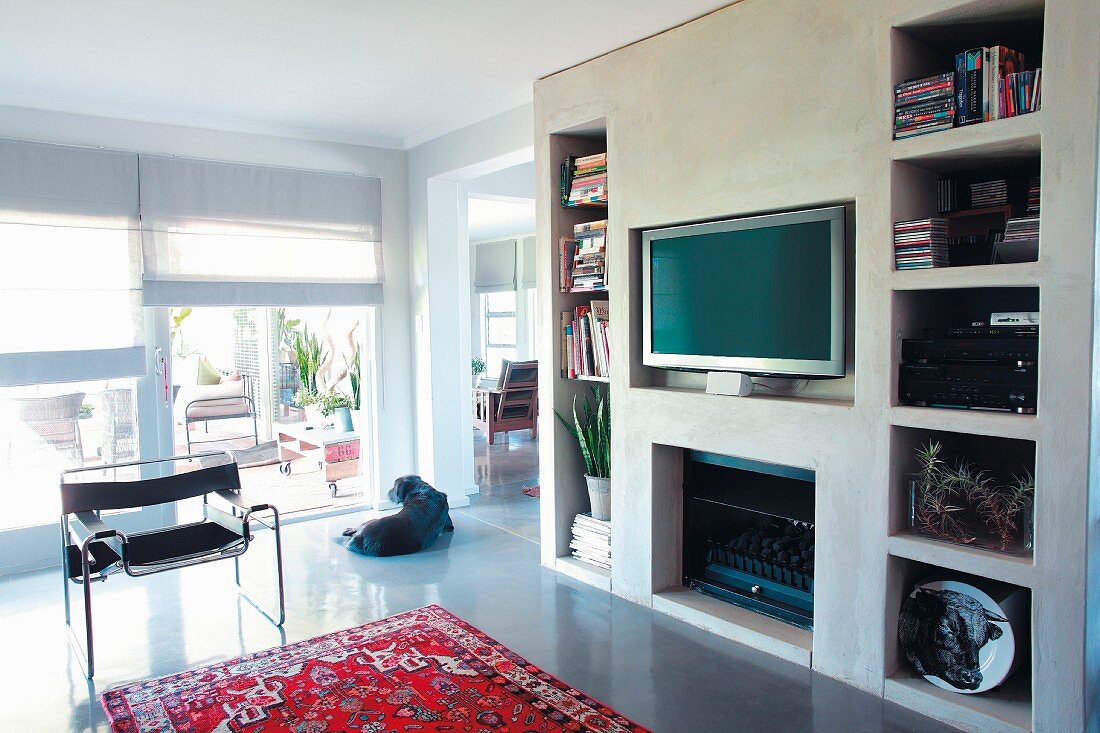 Betonierte Wandscheibe mit verschiedenen Aussparungen für Bücher, Fernseher und Gaskamin in offenem Wohnraum mit Blick auf Terrasse