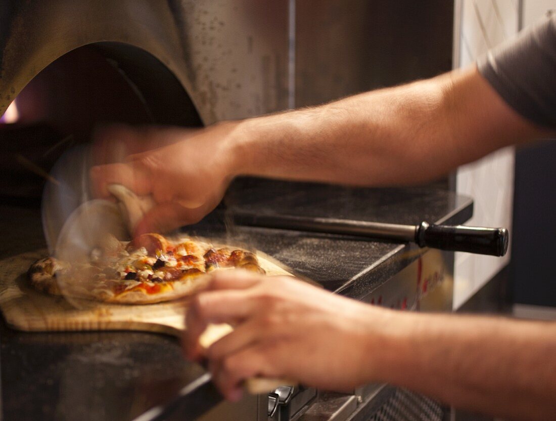 A pizza baker slicing a pizza baked in a wood-fired oven