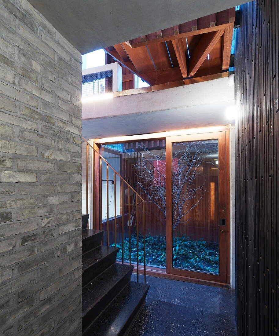 View from hall with black stone staircase and gallery into courtyard with bare tree in modern, architectural ambiance