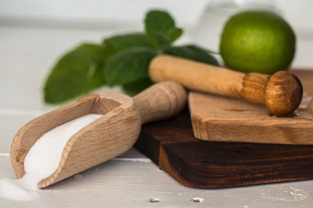 A muddler, limes, a wooden scoop with sugar on a wooden board