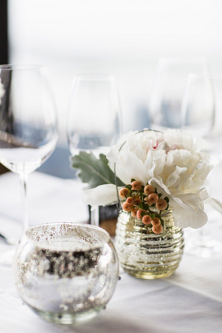 A silver tea light holder, a bunch of flowers and wine glasses on a table laid in white