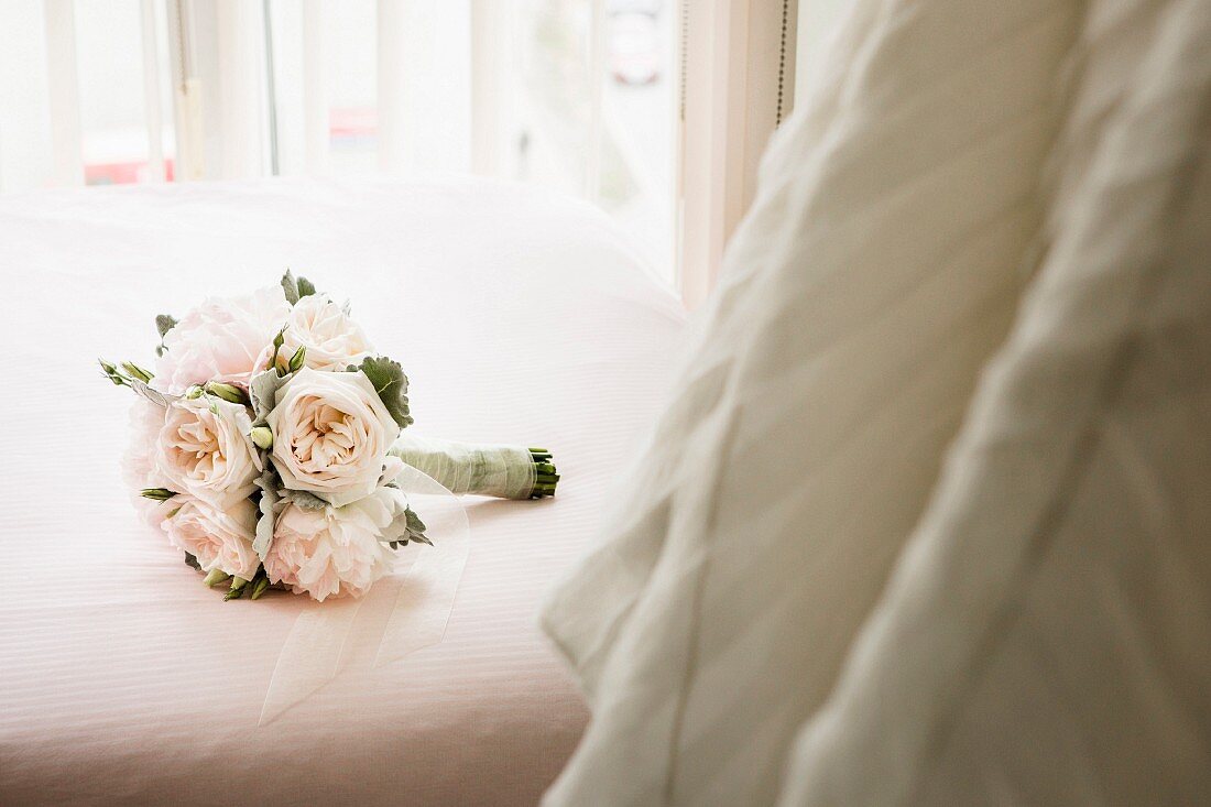 A bridal bouquet on a bed next to a hanging wedding dress