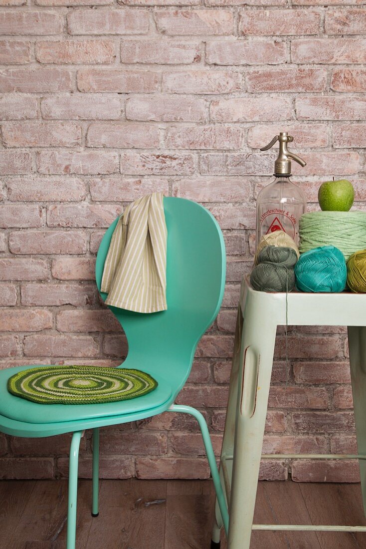 Crocheted seat pad on turquoise chair and crochet yarn and soda siphon on vintage stool against brick wall