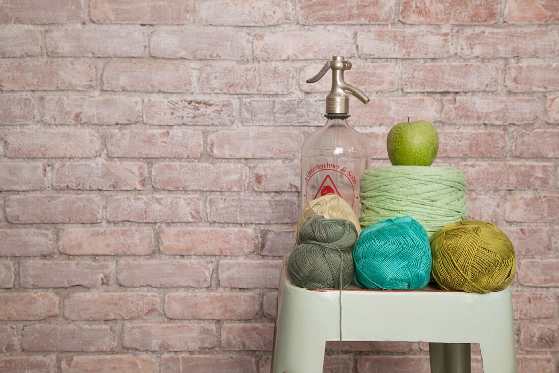 Crochet yarn in shades of green and blue and vintage soda siphon on vintage stool against brick wall