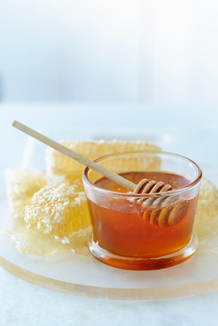 Honig mit Honiglöffel im Glasschälchen, dahinter Honigwaben