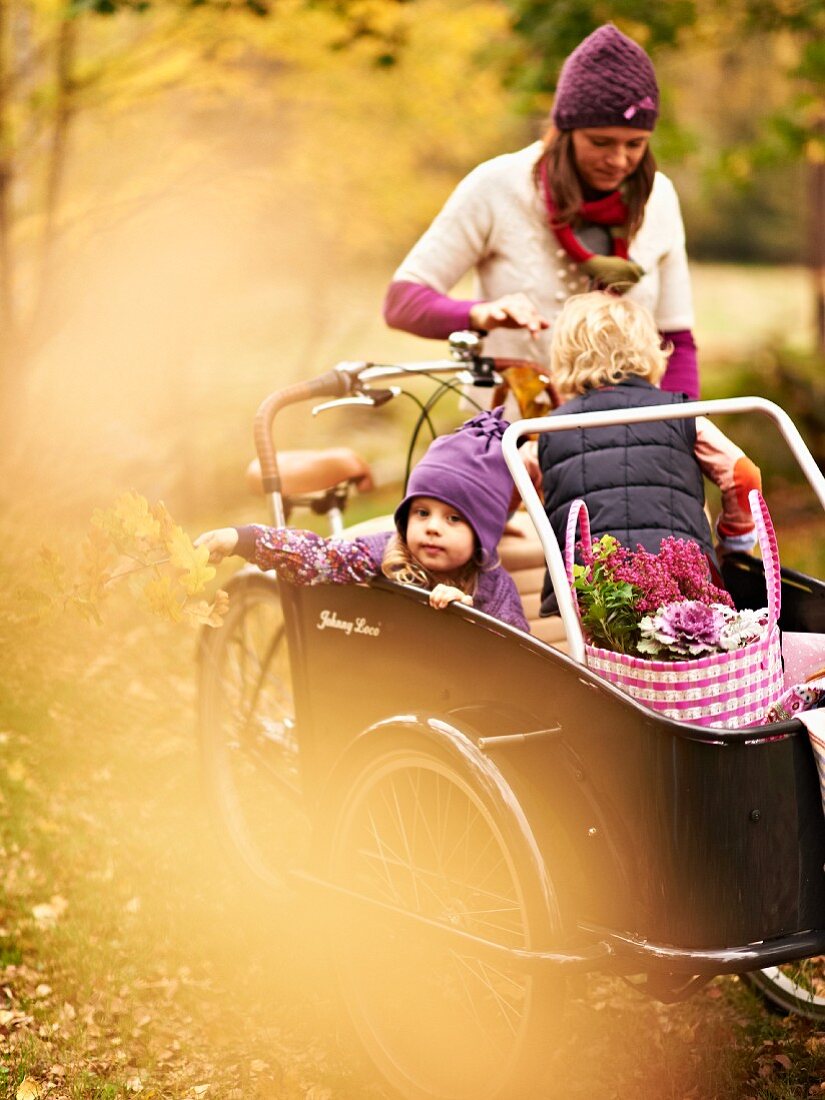 Mother pushing children and picnic baskets in box of cargo bike through autumn landscape