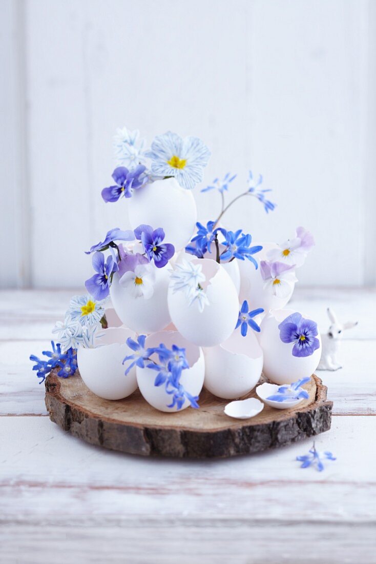 A pyramid of egg shells filled with blue spring flowers on a slice of bark