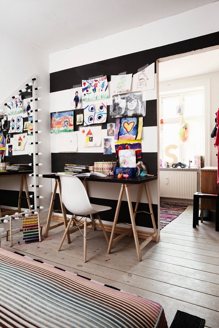 Open doorway next to white classic chair at desk below children's' drawings hung on black and white striped wall