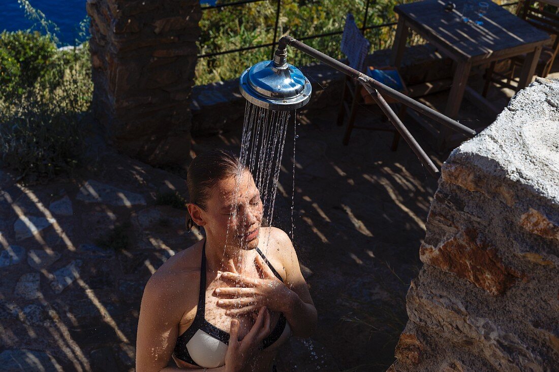 A woman wearing a bikini under a shower outside a rustic stone house