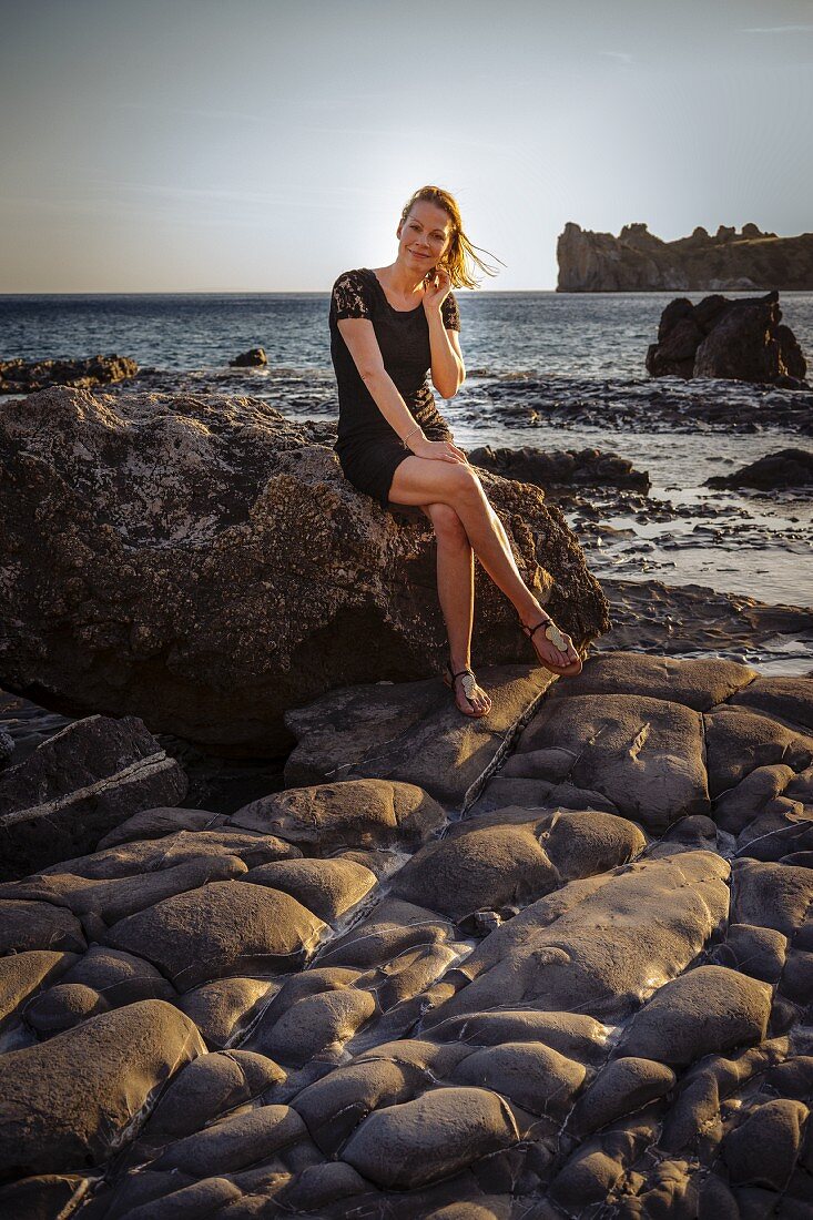Frau in schwarzem Minikleid sitzt auf Felsen am Meer