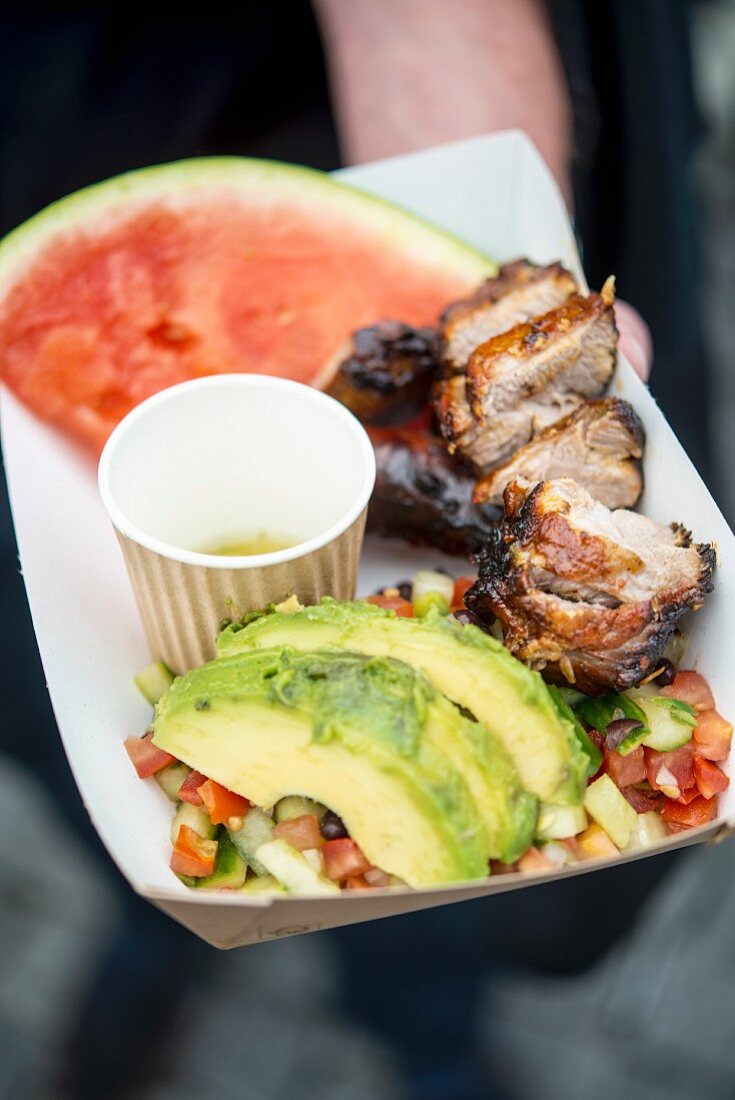 A hand holding grilled pork with a vegetable salad, avocado and melon in a paper dish