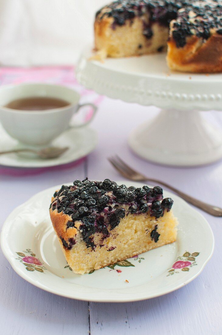 A slice of blueberry cake with icing sugar served with a cup of tea