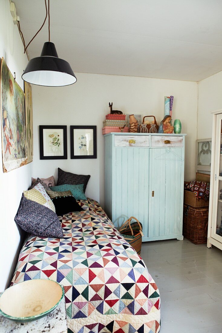 Colourful quilt and various scatter cushions on single bed next to pale blue wardrobe in simple bedroom