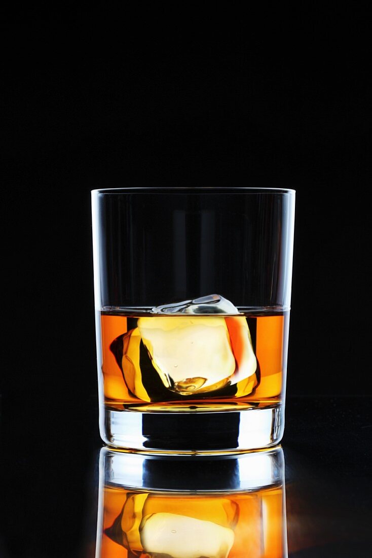 A glass of whiskey with an ice cube on a black surface