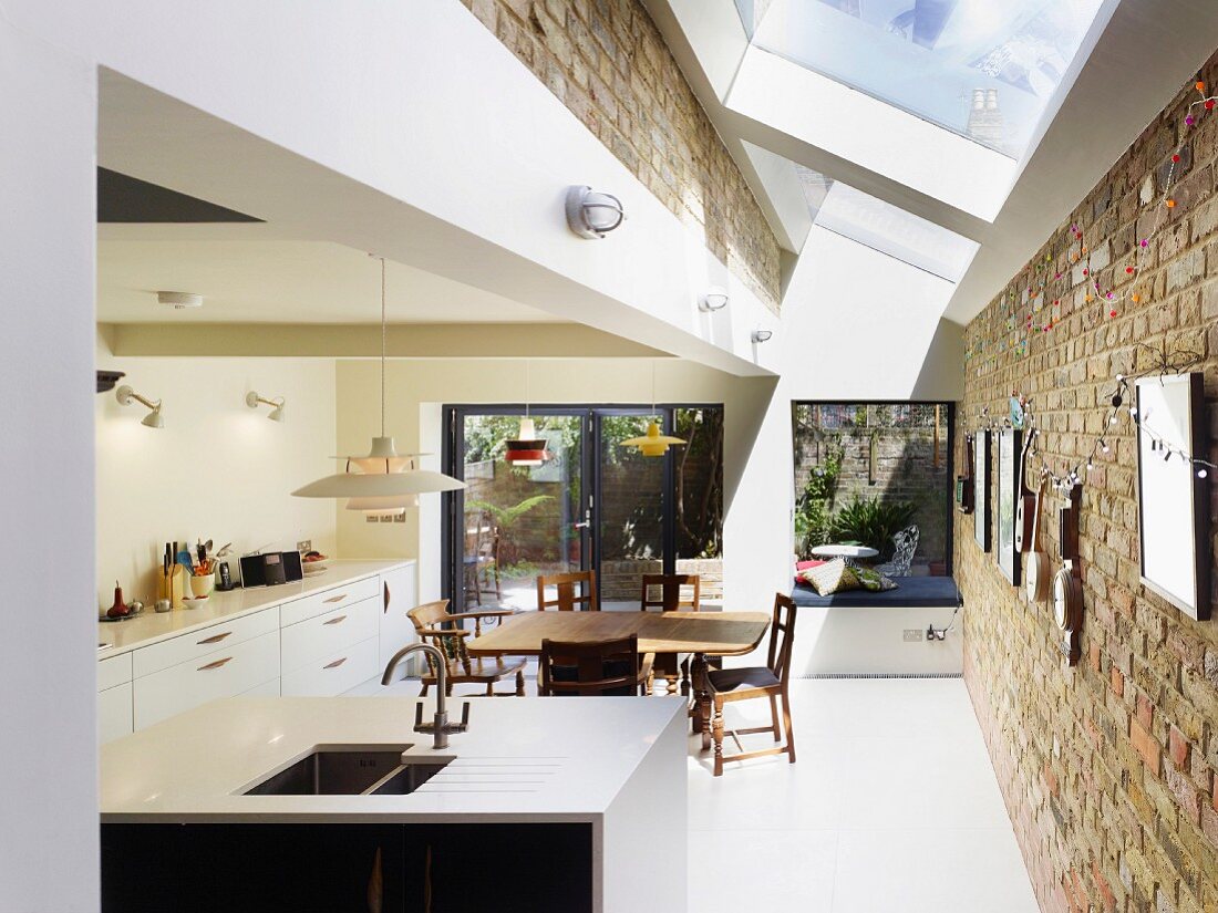 Strip of skylights with white frames and old brick walls in open-plan kitchen with dining area and view of seating area on summery terrace