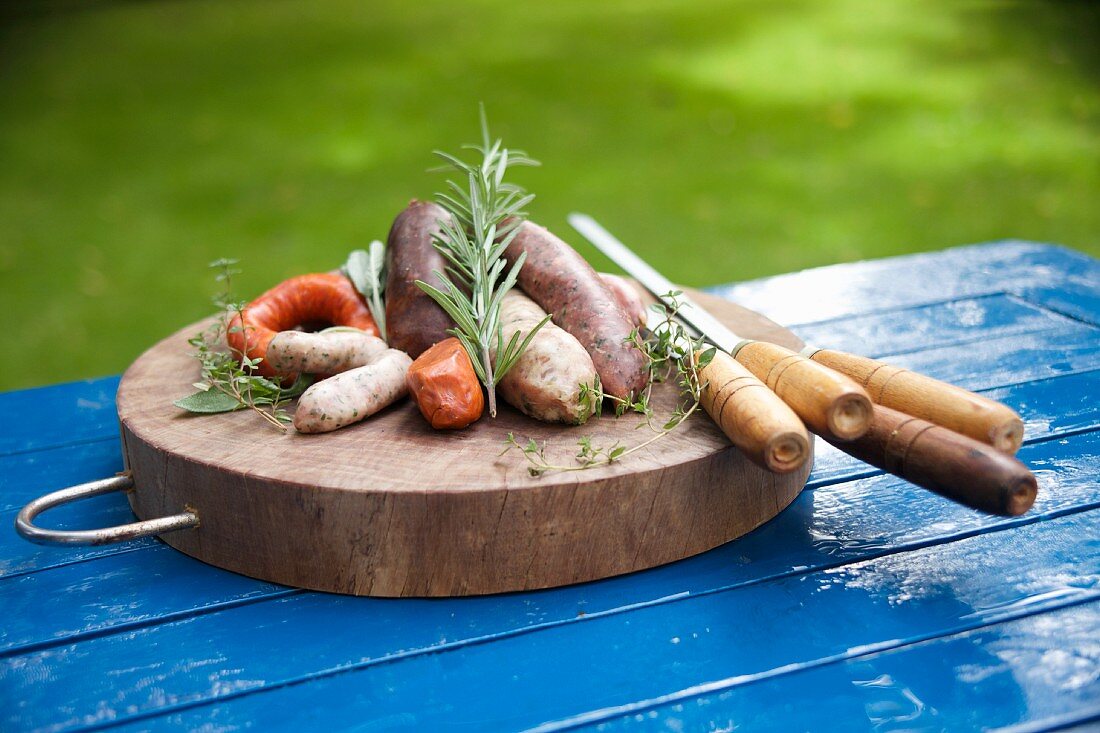 A wooden platter with various uncooked sausages, herbs and skewers