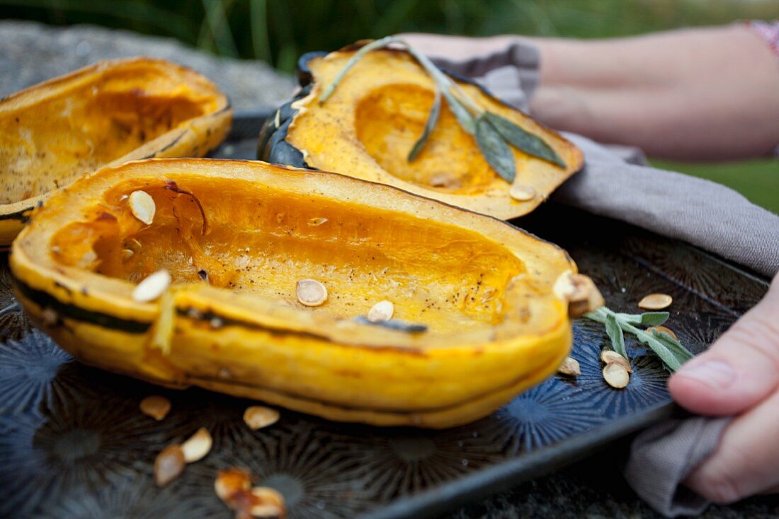 A woman holding a tray of roasted, hollowed out squash with seeds under Sage