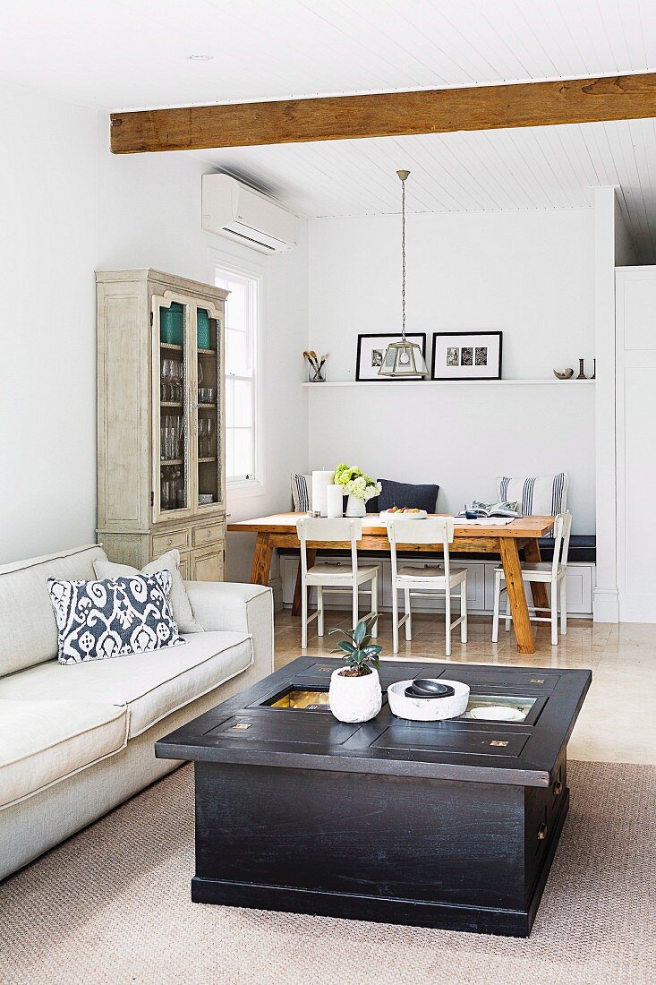 Box-shaped coffee table made of dark-colored wood with drawers and light sofa in an open living room, in the background dining area in the corner of the room