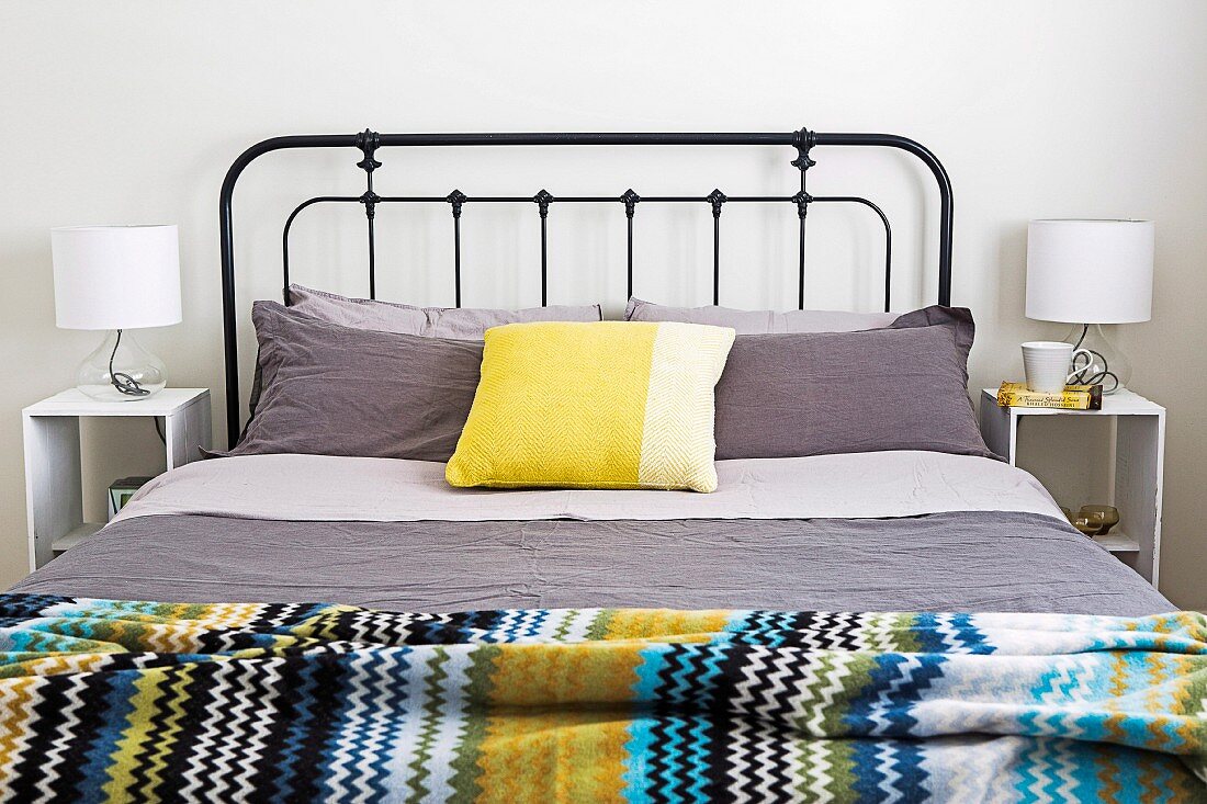 Doppelbett mit schwarzem Metallgittergestell, grauer Bettwäsche, Kissen in Gelbtönen und bunt gestreifter Tagesdecke
