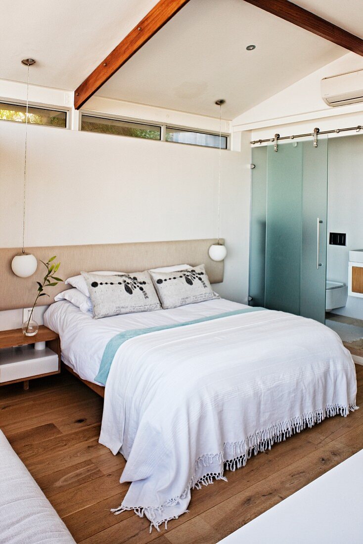 Double bed with white bedspread against wall with strip of narrow transom windows; ensuite bathroom to one side with half-open sliding glass door in modern bedroom