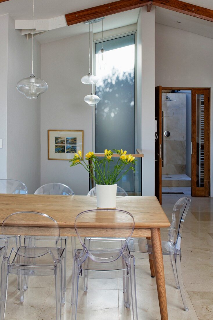 Vase of yellow freesias on wooden table, Ghost chairs and delicate pendant lamps with glass lampshades in modern interior