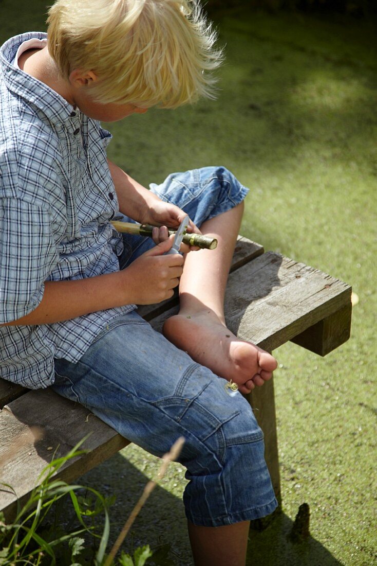 Blonde boy carving a wooden flute sitting on wooden jetty next to garden pond