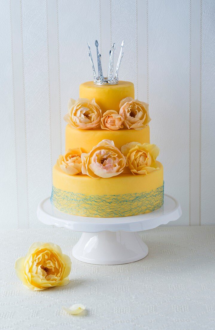 A three-tier, yellow fondant cake decorated with rose petals and a crown