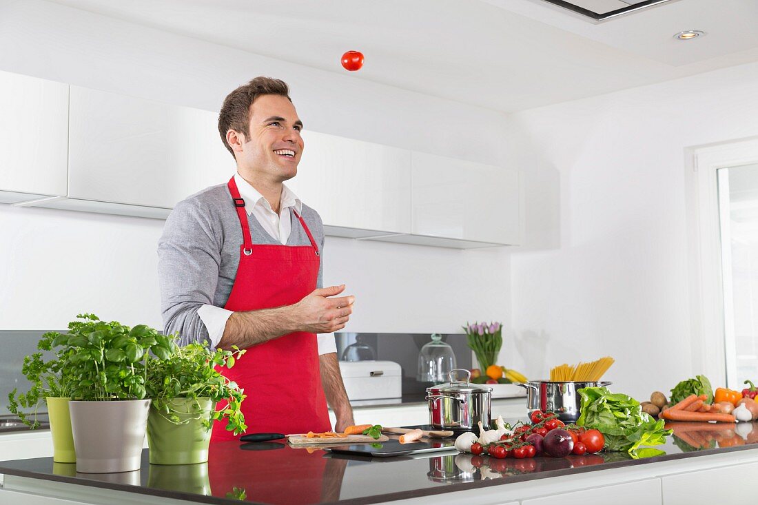 A man wearing an apron laughing and juggling tomatoes in a kitchen