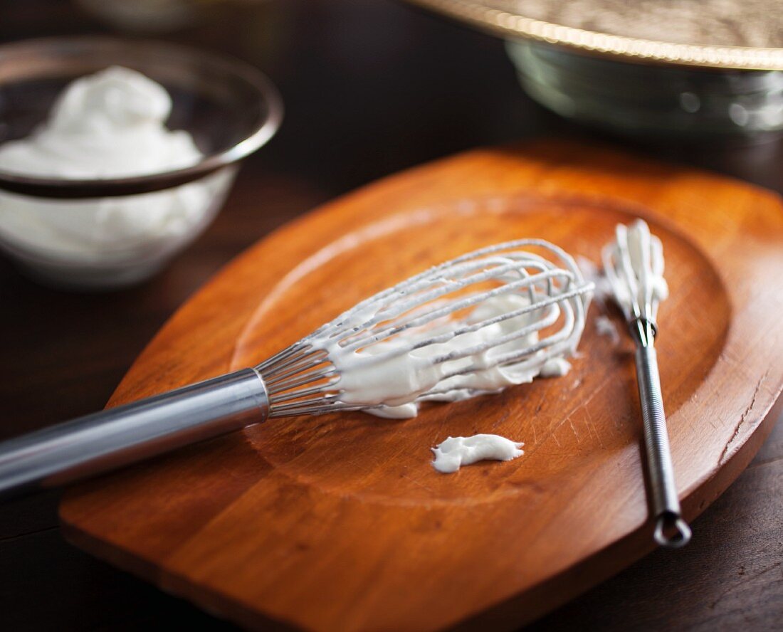 Two whisks with freshly whipped cream on a wooden board