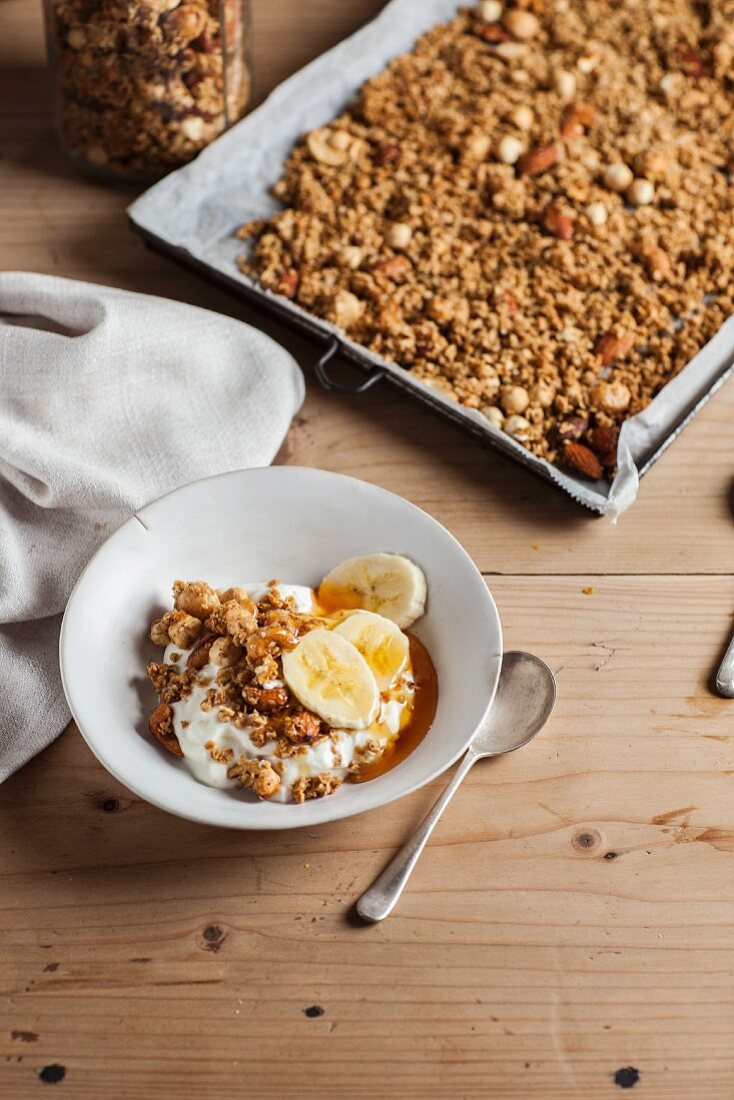 Granola with nuts and honey on a baking tray with a bowl of yoghurt muesli and banana in the foreground