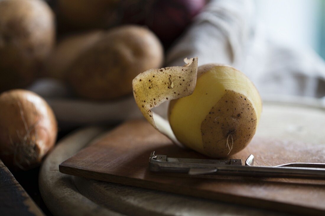 Kartoffel und Sparschäler auf Holzbrett, im Hintergrund Kartoffeln und Zwiebeln