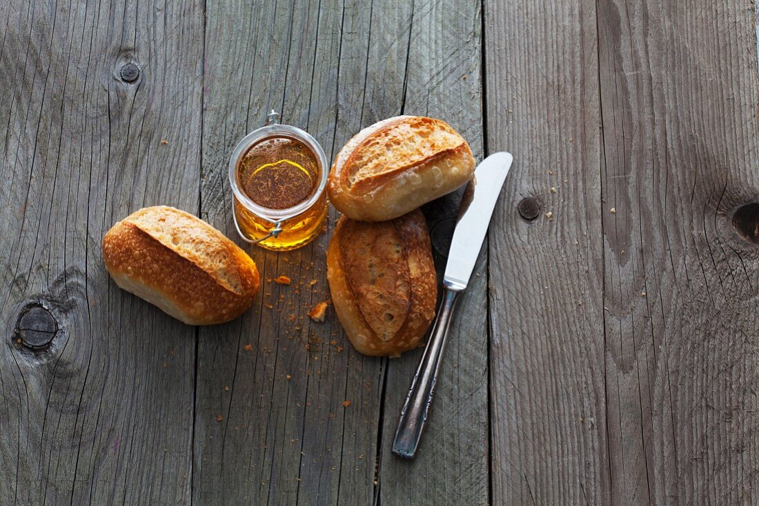 Three rolls, a knife and a jar of honey on a wooden surface